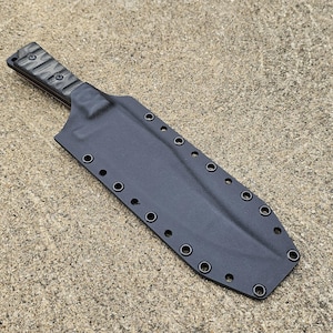 Könnte beinhalten: Ein schwarzes Messer mit strukturiertem Griff und passender Scheide. Die Scheide hat mehrere Ösen an den Rändern. Das Messer und die Scheide liegen auf einer strukturierten, hellbraunen Oberfläche.