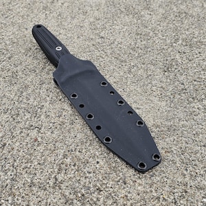 Puede incluir: Un cuchillo negro con una funda. El cuchillo tiene una hoja puntiaguda y un mango texturizado. La funda es negra con múltiples ojales a lo largo de los bordes. El cuchillo y la funda están sobre una superficie gris texturizada.