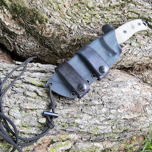 Puede incluir: Una funda de cuchillo de cuero negro con una capa exterior de tela gris oscuro y un cordón de paracord negro. La funda tiene tres botones a presión negros y está diseñada para sujetar un cuchillo de hoja fija.