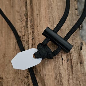 以下が含まれることがあります： 黒いパラコードのランヤードに、黒い木製のトグルと白いタグが付いています。