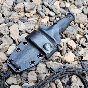 Könnte beinhalten: Ein schwarzes Messer mit einer schwarzen Scheide und einer schwarzen Schnur. Das Messer hat einen strukturierten Griff und eine schwarze Klinge. Die Scheide hat einen Druckknopfverschluss und eine Schlaufe zum Befestigen der Schnur.