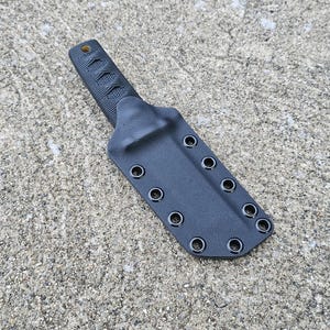 Puede incluir: Un cuchillo negro con un mango texturizado y una funda a juego. La funda tiene múltiples ojales a lo largo de los bordes. El cuchillo y la funda descansan sobre una superficie gris texturizada.