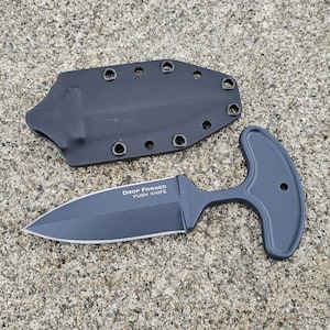 Könnte beinhalten: Ein schwarzes, geschmiedetes Push-Messer mit einem T-förmigen Griff und einer passenden Scheide. Das Messer hat eine doppelschneidige Klinge und der Text "DROP FORGED PUSH KNIFE" ist auf der Klinge sichtbar. Die Scheide hat mehrere Löcher.