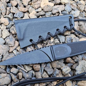 May include: A black fixed-blade knife with a serrated edge and a textured handle featuring a circular spiral design. The knife includes a matching black sheath with a woven cord lacing detail and a long black cord lanyard attached to the handle.