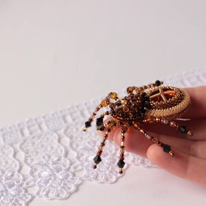 May include: A beaded spider brooch with a gold and brown body and black accents. The spider is sitting on a hand.