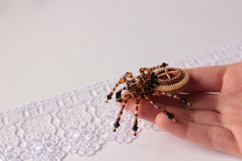 May include: A beaded spider brooch with a gold and brown body and black accents. The spider is sitting on a hand.