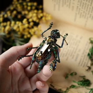 May include: A green and gold beaded insect brooch with a gold pin. The brooch is held in a hand against a book.