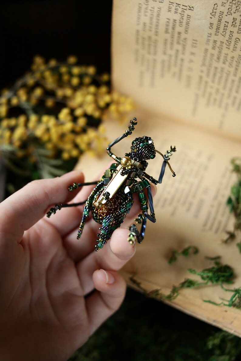 May include: A green and gold beaded insect brooch with a gold pin. The brooch is held in a hand against a book.