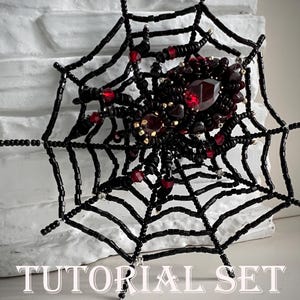 May include: A beaded spider and web design. The spider is black with red accents and a large red gem at its center. The web is black and the text "TUTORIAL SET" is at the bottom.