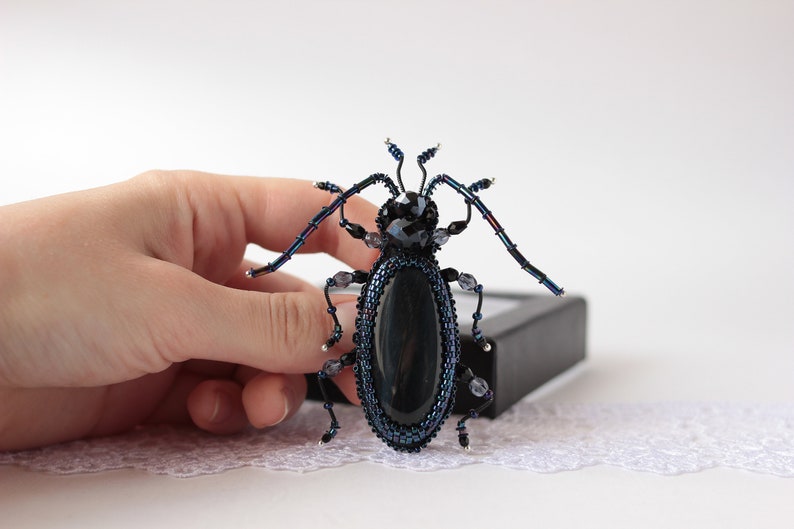 May include: A black and blue beaded beetle brooch with iridescent wings and a black oval stone in the center.