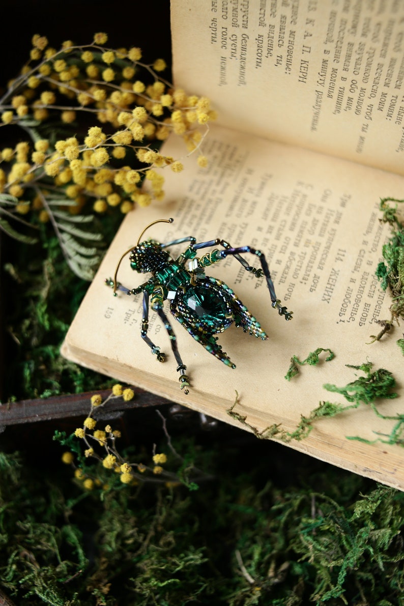 May include: A green and blue beaded insect brooch with a large emerald green gemstone in the center. The brooch is sitting on an open book with yellow flowers and green moss.