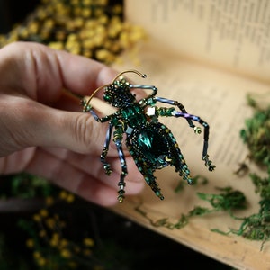 May include: A green and gold beaded insect brooch with a large emerald green gemstone in the center. The brooch is held in a hand against a background of a book and moss.