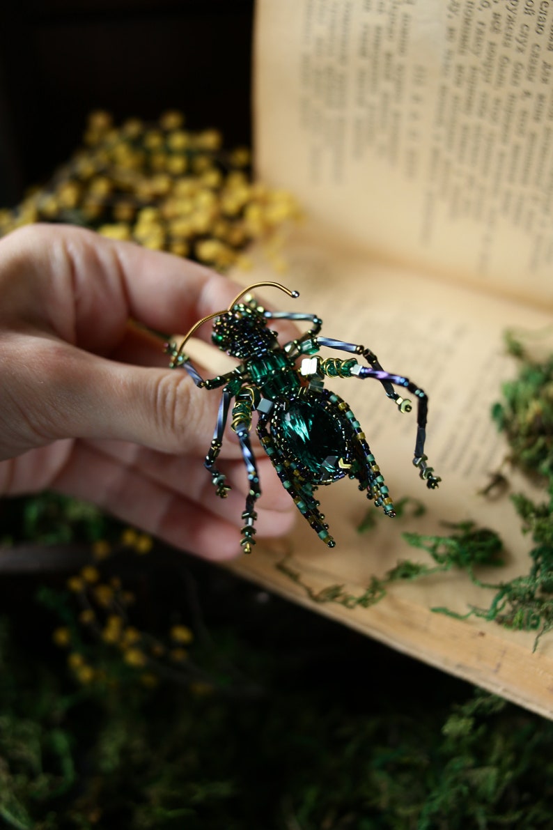 May include: A green and gold beaded insect brooch with a large emerald green gemstone in the center. The brooch is held in a hand against a background of a book and moss.