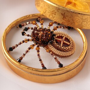 May include: A beaded spider brooch with a brown and gold body and black beads for legs. The brooch is displayed in a gold box.