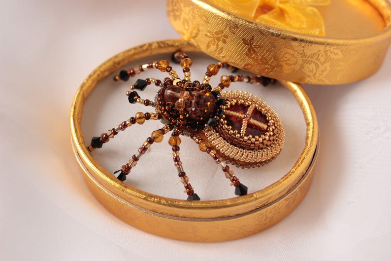 May include: A beaded spider brooch with a brown and gold body and black beads for legs. The brooch is displayed in a gold box.