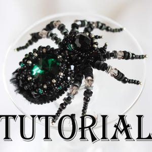 以下が含まれることがあります： 大きな緑色の宝石と黒いビーズが付いた、ビーズで作られたクモのブローチ。クモには複数の脚があり、透明な表面に表示されています。「TUTORIAL」という単語は、黒色の様式化されたテキストで書かれています。