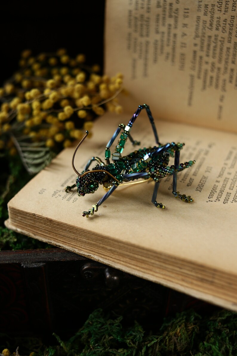 May include: A green and blue beaded grasshopper brooch with iridescent accents. The brooch is sitting on an open book with yellow flowers in the background.