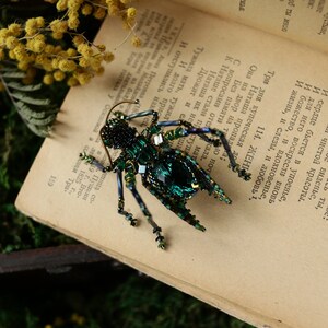 May include: A green and black beaded insect brooch with a large emerald green gem in the center. The brooch is sitting on a vintage book.