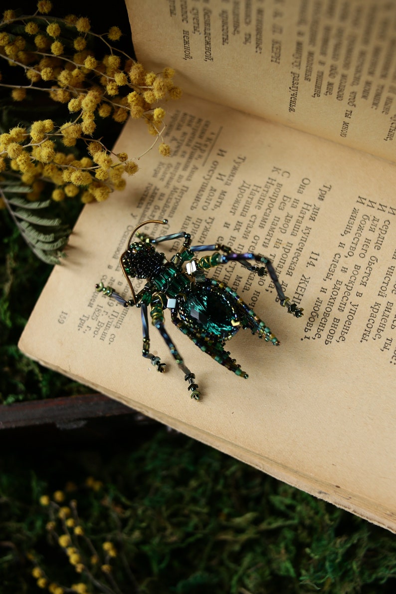 May include: A green and black beaded insect brooch with a large emerald green gem in the center. The brooch is sitting on a vintage book.