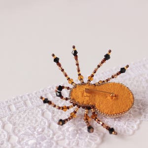 May include: A beaded spider brooch with a yellow felt body and black and brown beads. The brooch is on a white lace background.