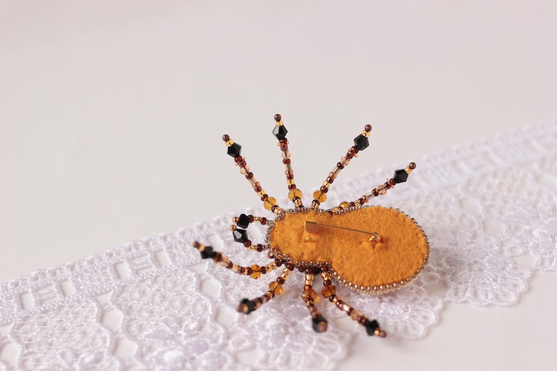 May include: A beaded spider brooch with a yellow felt body and black and brown beads. The brooch is on a white lace background.