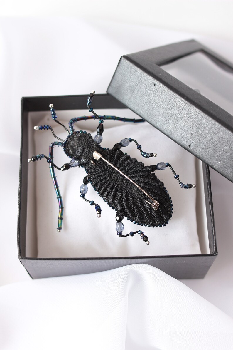 May include: A black beaded beetle brooch with iridescent blue and black accents. The brooch is in a black box with a clear lid.