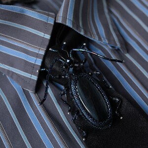 May include: A black and blue beaded beetle brooch with iridescent accents. The brooch is pinned to a striped blue and gray shirt.