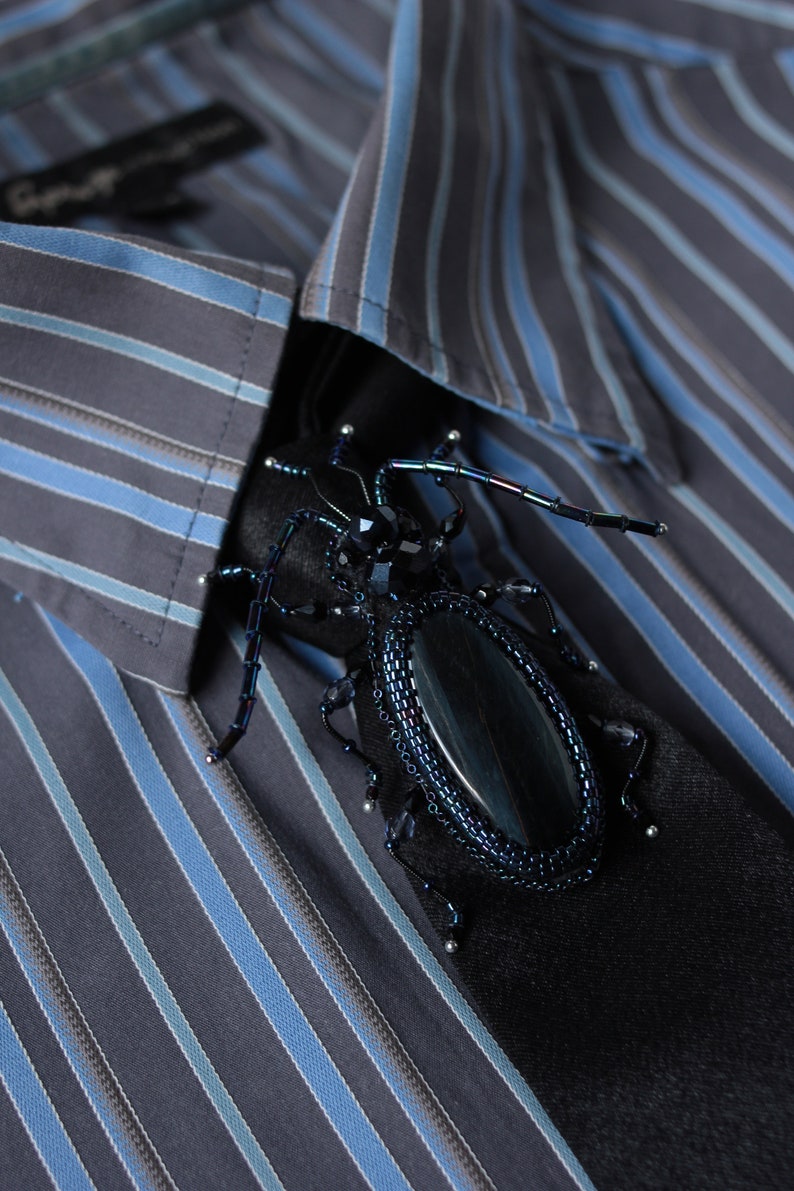 May include: A black and blue beaded beetle brooch with iridescent accents. The brooch is pinned to a striped blue and gray shirt.
