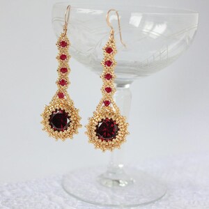 Peut inclure: Une paire de longues boucles d'oreilles dorées avec des accents de pierres précieuses rouges. Les boucles d'oreilles présentent une grande pierre précieuse rouge ronde entourée de perles dorées.