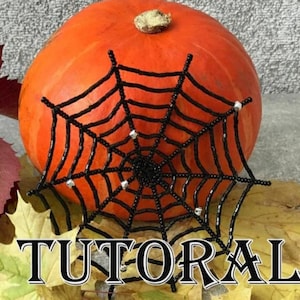 May include: A decorative orange pumpkin with a black beaded spiderweb. The spiderweb features silver embellishments and is positioned in front of the pumpkin. The word "TUTORAL" is visible at the bottom of the image.