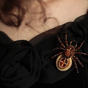 May include: A black beaded spider brooch with a gold and amber centre. The brooch is pinned to a black fabric background.