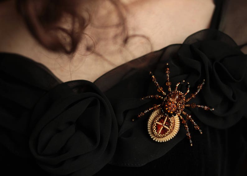 May include: A black beaded spider brooch with a gold and amber centre. The brooch is pinned to a black fabric background.