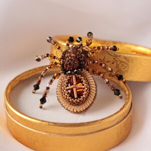 May include: A beaded spider brooch with a brown and gold body and black legs. The spider is sitting in a small, round, gold box.