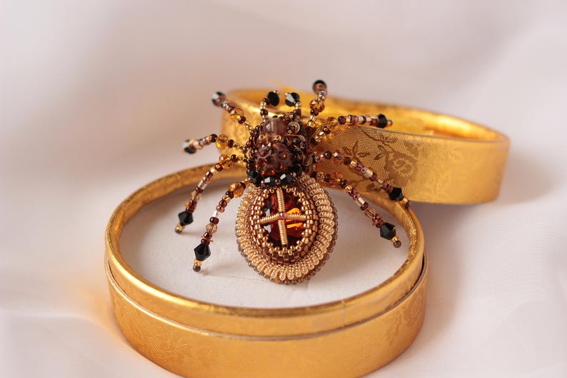 May include: A beaded spider brooch with a brown and gold body and black legs. The spider is sitting in a small, round, gold box.