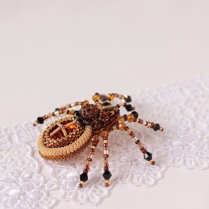 May include: A beaded spider brooch with a brown and gold body and black legs. The spider is sitting on a white lace background.