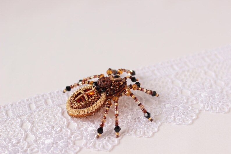May include: A beaded spider brooch with a brown and gold body and black legs. The spider is sitting on a white lace background.