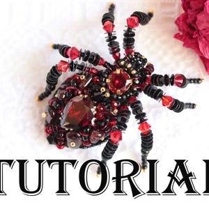 Peut inclure: Une broche araignée perlée noire, rouge et dorée. L'araignée présente une grosse pierre rouge facettée au centre et est entourée de petites perles. Le mot "TUTORIAL" est écrit en lettres noires stylisées en dessous.