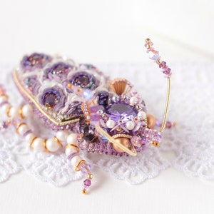May include: A purple and gold beaded insect brooch with a large amethyst gemstone. The brooch is on a white lace background.