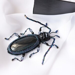 May include: A beaded beetle brooch with a black gemstone cabochon and iridescent blue and black beads. The beetle is set on a white fabric background.
