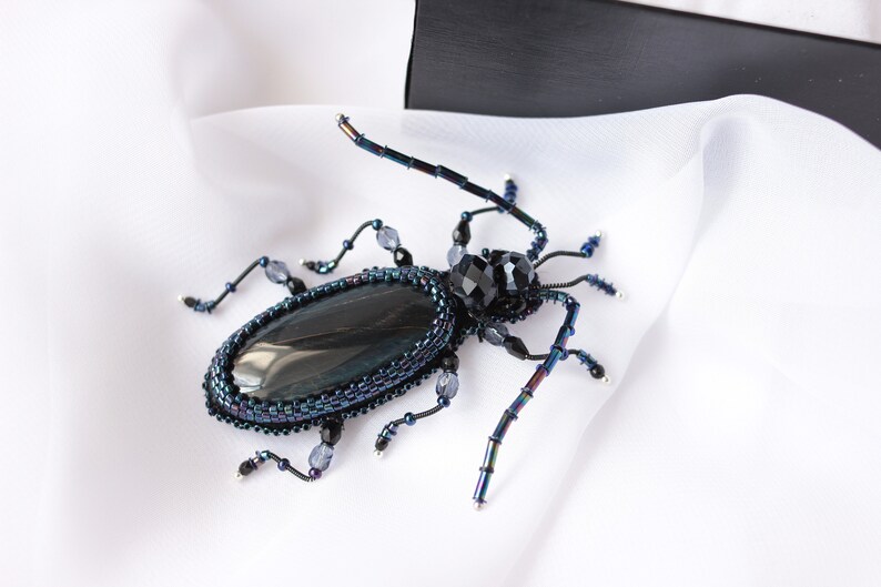 May include: A beaded beetle brooch with a black gemstone cabochon and iridescent blue and black beads. The beetle is set on a white fabric background.