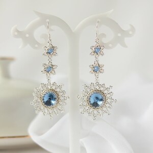 Peut inclure: Boucles d'oreilles pendantes argentées avec des accents de cristal bleu. Les boucles d'oreilles présentent trois étoiles en cristal et un plus grand cristal rond.