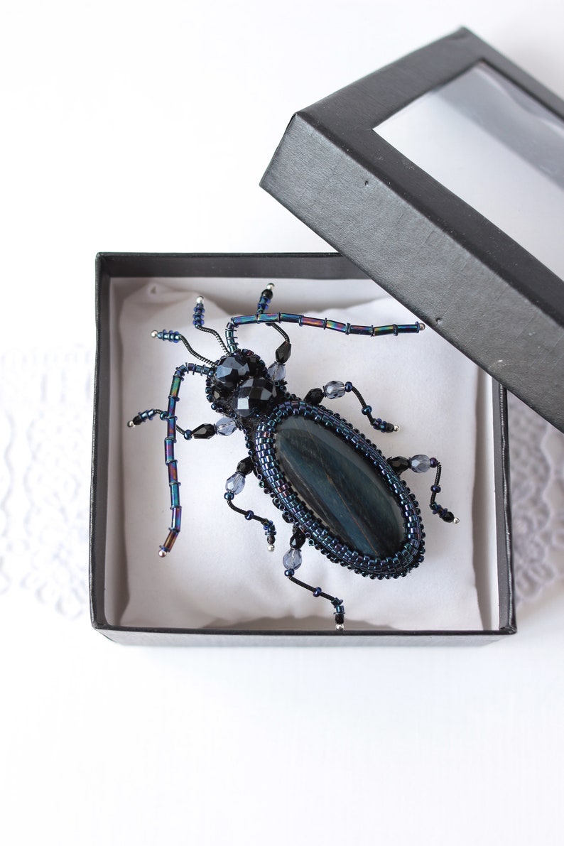 May include: A black and blue beaded beetle brooch with iridescent wings and a dark blue gemstone body. The brooch is displayed in a black box with a clear lid.