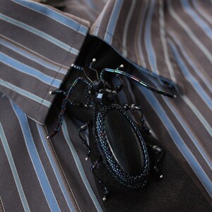 May include: A black beaded insect brooch with a black stone body and iridescent wings. The brooch is pinned to a striped shirt.