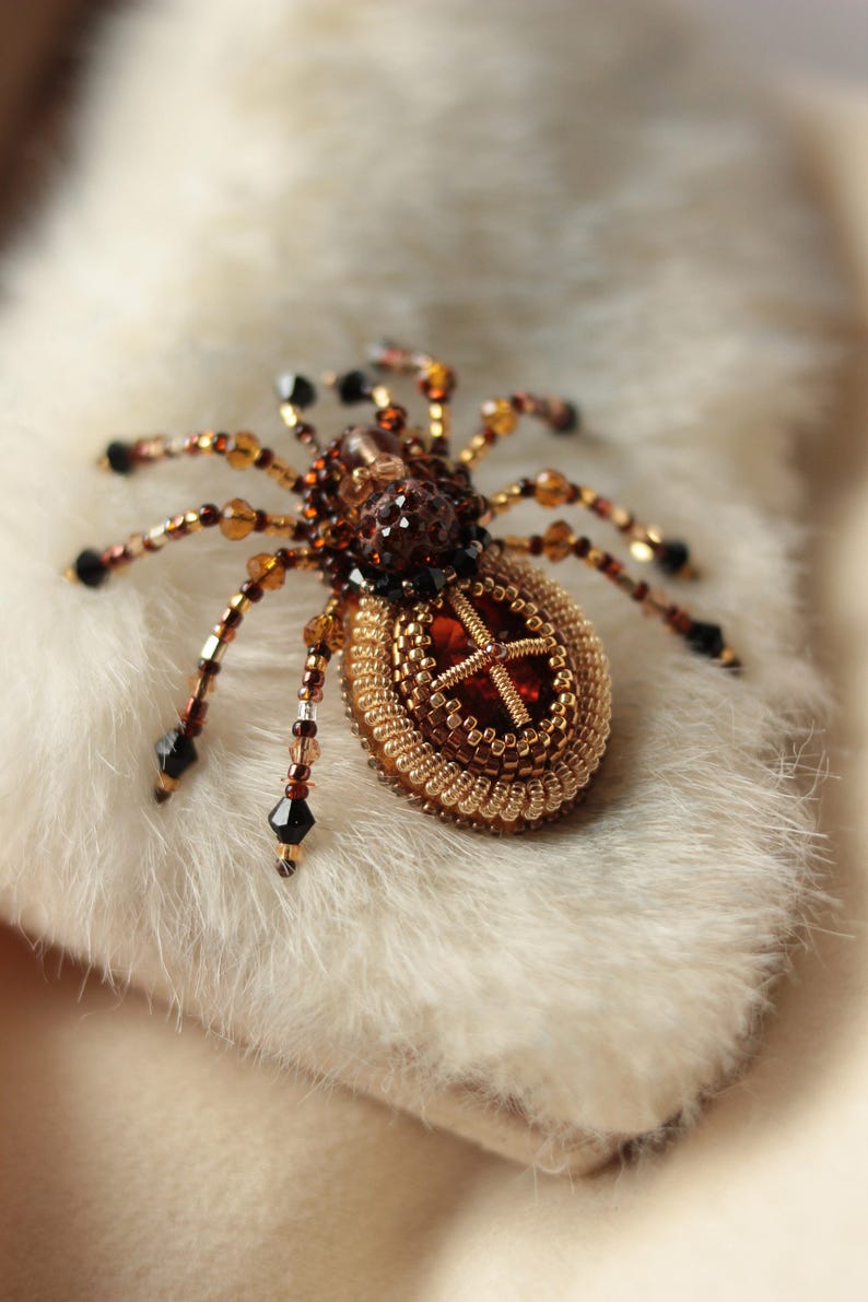 May include: A beaded spider brooch with a brown and gold body and black legs. The spider is sitting on a white fur surface.