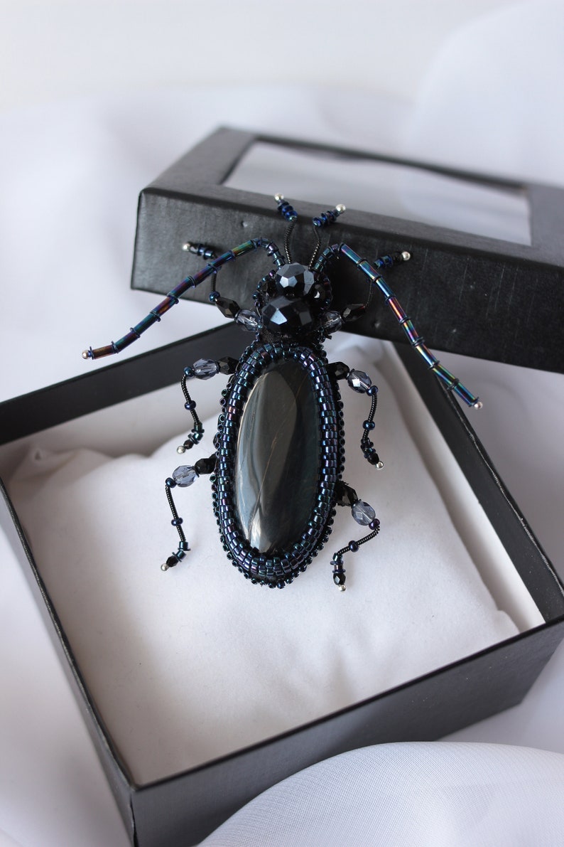 May include: A black beaded beetle brooch with iridescent wings and a black gemstone body. The brooch is displayed in a black box with a white interior.