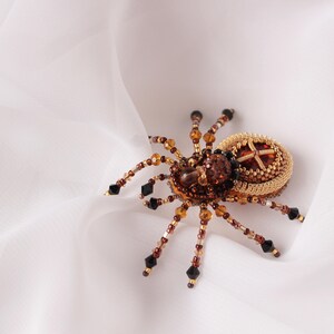 May include: A beaded spider brooch in gold and brown tones. The spider has an intricate body with a cross-shaped design at its centre.