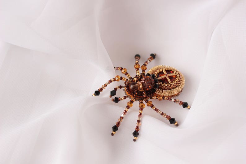 May include: A beaded spider brooch in gold and brown tones. The spider has an intricate body with a cross-shaped design at its centre.