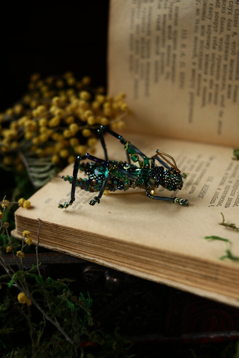 May include: A green and black beaded grasshopper brooch with gold wire accents. The brooch is sitting on an open book with yellow flowers in the background.