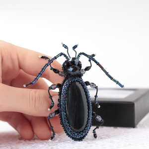 May include: A beaded beetle brooch with a black gemstone body and iridescent blue wings. The brooch is held in a hand against a white background.