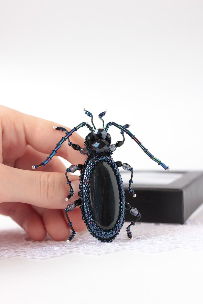 May include: A beaded beetle brooch with a black gemstone body and iridescent blue wings. The brooch is held in a hand against a white background.